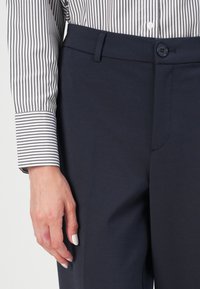Gros plan d'une personne portant une chemise blanche et noire à rayures verticales, rentrée dans un pantalon formel bleu marine avec un bouton et des passants de ceinture.
