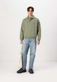 Sudadera verde con cuello, con un logo en el pecho, combinada con jeans azul claro y zapatillas negras. Se sostiene una bolsa de tela en una mano.