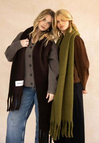 Deux femmes portant des écharpes : l'une marron foncé avec des franges, l'autre vert olive. Les deux écharpes ont une texture douce et sont larges, tombant sur leurs tenues.