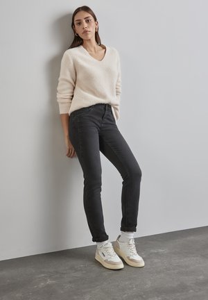 Pull en tricot beige clair avec un col en V ; jeans noirs slim ; baskets blanches et beiges ; le mannequin pose devant un fond neutre.