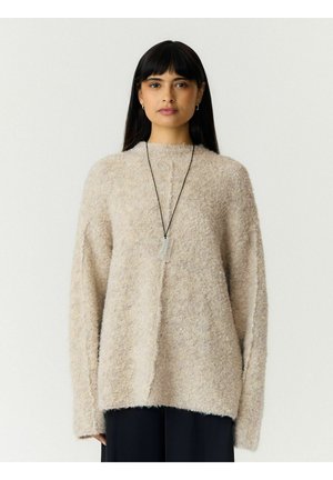 Femme aux longs cheveux noirs portant un pull beige duveteux et un pantalon noir, avec un pendentif peigne transparent sur un collier en cordon noir.