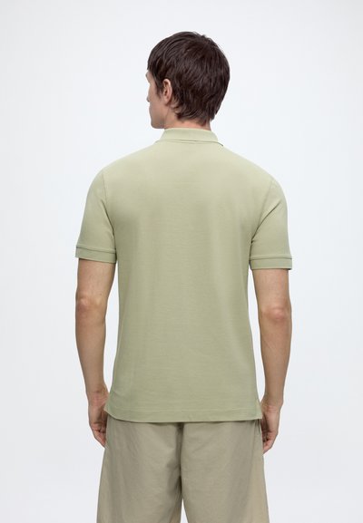 Homme portant un polo vert clair à manches courtes et un short beige, debout, de dos sur un fond blanc uni.