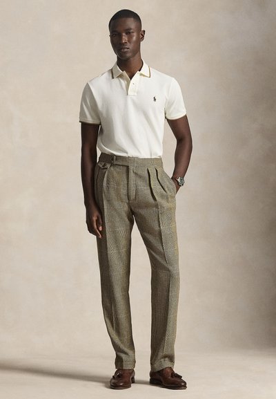 Vyras, dėvintis baltą trumpomis rankovėmis polo marškinėlius, rudus languotus aukšto liemens kelnes, rudus tasselių tipo batelius ir rankinį laikrodį, stovintis prieš neutralią foną.