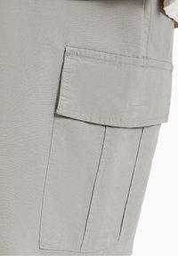 Ljusgrått tyg med en fin struktur, utrustat med en stor patch-ficka på sidan, sydd med en kontrasterande tråd.