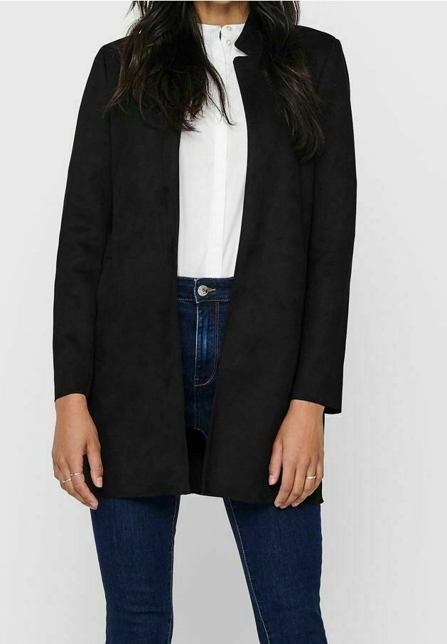 Vrouw in een zwarte open voorkant lange mouw jasje over een witte blouse met knopen en donkerblauwe jeans, staand tegen een effen achtergrond.