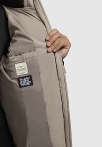 Beige gefütterte Jacke mit glatter, glänzender Textur. Besitzt ein eingenähtes Etikett und ein schwarzes Logo-Patch, von einer Hand gehalten.