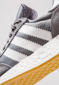 Grå idrottssko med vita zick-zack-ränder, övre del i texturerad tyg, gummisul med hexagonmönster, vadderad krage.