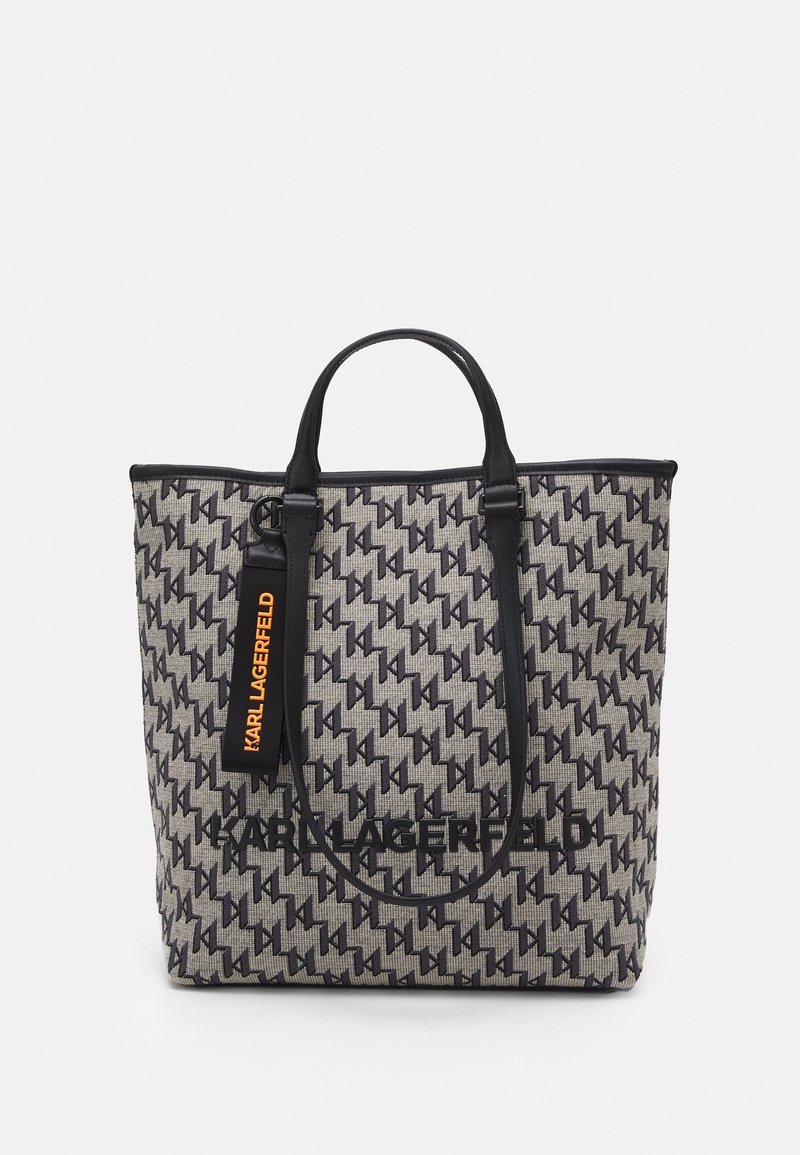 Bolsa tote en tela gris texturizada con un patrón geométrico negro, con asas de cuero negro y un logo negro prominente, además de una etiqueta naranja.