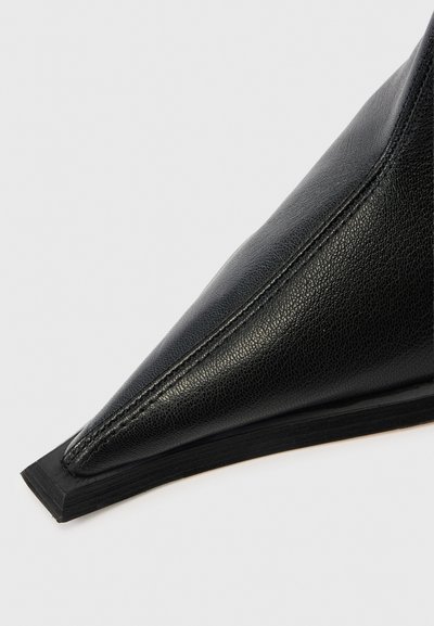 Μαύρο δερμάτινο παπούτσι με μυτερή μύτη, που διαθέτει υφή στην επιφάνεια και διακριτική ραφή. Η σόλα είναι επίπεδη με ελαφρώς ανυψωμένη ελαστική άκρη.