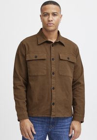Chaqueta de botones marrón hecha de algodón, con dos bolsillos en el pecho con botones negros, un cuello puntiagudo y mangas largas. La lleva puesta sobre unos vaqueros de mezclilla azules.