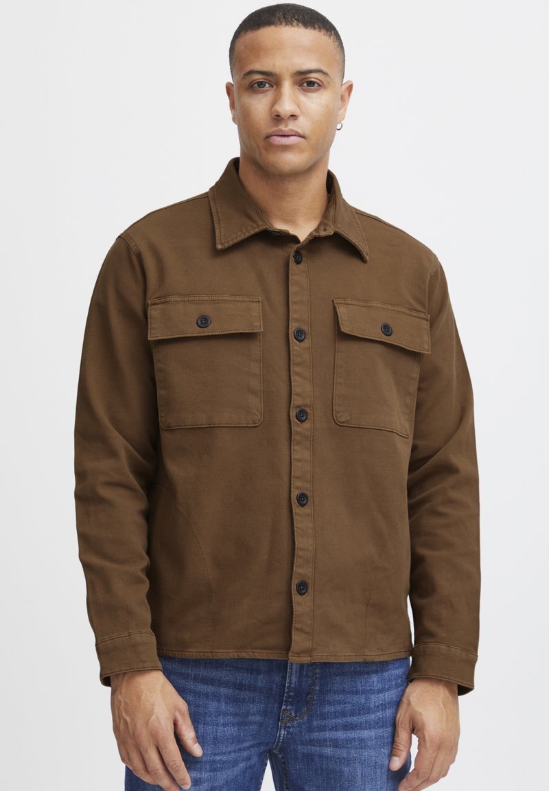 Chaqueta de botones marrón hecha de algodón, con dos bolsillos en el pecho con botones negros, un cuello puntiagudo y mangas largas. La lleva puesta sobre unos vaqueros de mezclilla azules.