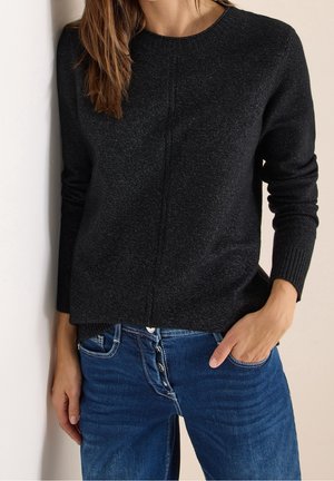 Femme portant un pull à manches longues gris foncé et un jean bleu avec boutons apparents, une main dans la poche, sur un fond clair.