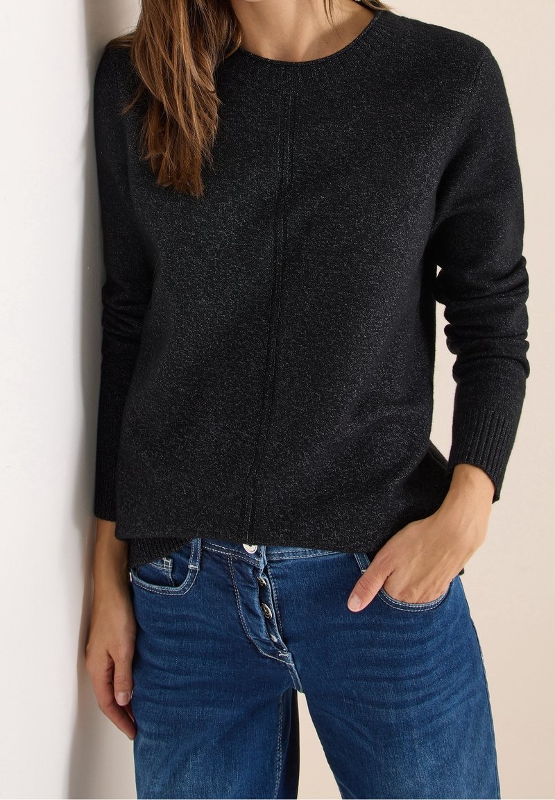 Femme portant un pull à manches longues gris foncé et un jean bleu avec boutons apparents, une main dans la poche, sur un fond clair.