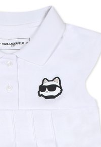 Polo shirt en coton blanc à manches courtes, avec un badge représentant un chat noir et blanc portant des lunettes de soleil sur le côté gauche de la poitrine.