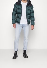 Grön puffjacka med svarta accenter, framdragkedja och huva, bärs över en vit t-shirt och ljusblå jeans. Svarta ankelskålar.