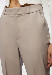 Beżowe, wysokie spodnie o gładkiej fakturze, z bocznymi kieszeniami i prostym krojem. Posiadają minimalne przeszycia i płaski przód.