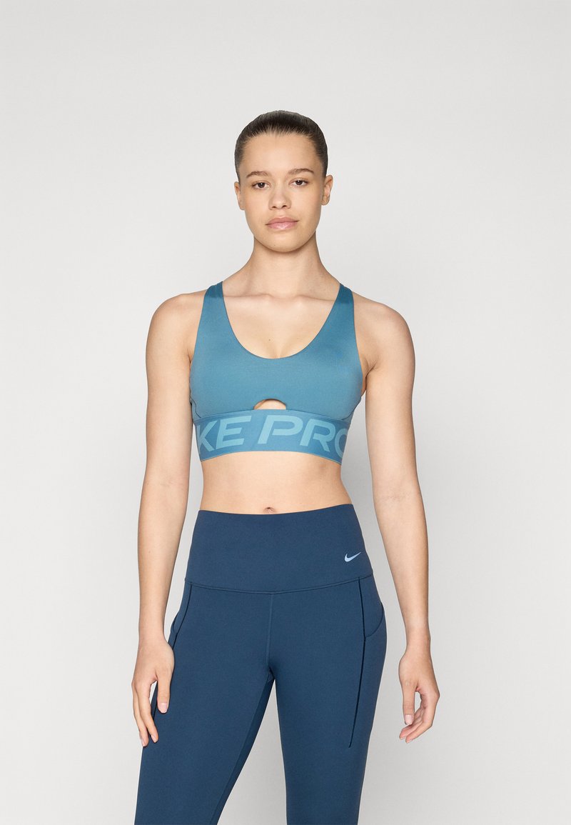 Modra športna modrčka z izrezom in širokim pasom, na katerem je napis "NIKE PRO" v svetlo modri barvi. Ujemajoča se s temno modrimi hlačami z visokim pasom.