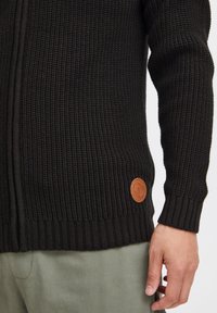 Schwarzer Strickcardigan mit strukturierter Oberfläche, der über gerippte Bündchen und einen gerippten Saum verfügt. Akzentuiert durch ein braunes Lederlabel nahe dem unteren Ende.