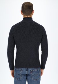 Dark gray ribbed sweater with a high collar and long sleeves, made of textured fabric. Paired with blue denim jeans.