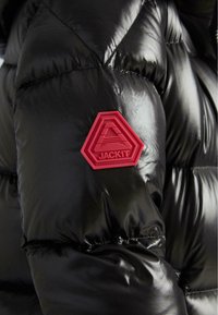 Giacca a vento nera con finitura lucida, cuciture in evidenza e una toppa esagonale rossa con "JACK1T" in rilievo.