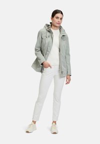Light grey, hooded jacket with zip closure, featuring a textured finish and pockets. Worn over a white sweater and paired with white pants.