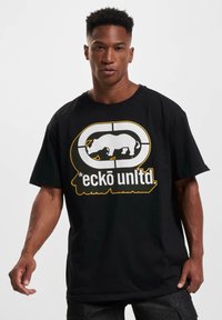 Juodas medvilninis marškinėliai su baltu raganosio grafika ir tekstu "eckō unlted." gelsva spalva. Trumpi rankoviai ir laisvas kirpimas. Paprastas, drąsus dizainas.