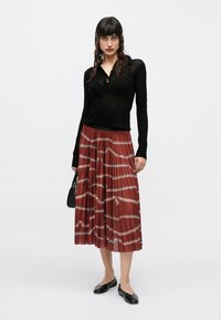 Haut noir à manches longues et col boutonné, ajusté. Jupe plissée bordeaux avec motifs géométriques blancs. Le mannequin porte des ballerines noires et tient un sac.