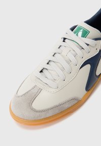Valge nahast sneaker hallide sametist aktsentidega, sinise logoga ja hallide paeladega, mis omab kummist kummipõhja ja tikandiga detaile.