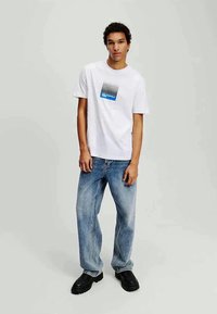 White cotton t-shirt featuring a blue and black striped graphic design. Paired with loose-fitting, light blue denim jeans and black shoes.