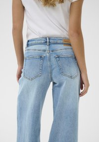 Person wearing light blue high-waisted wide-leg jeans and a tucked white sleeveless top, shown from back with hands relaxed by sides.