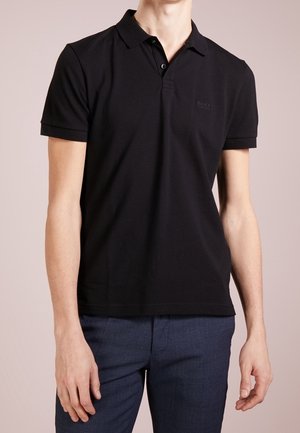 Mężczyzna ubrany w dopasowaną czarną koszulkę polo z krótkim rękawem i dwoma guzikami oraz ciemnoniebieskie spodnie na jednolitym tle.