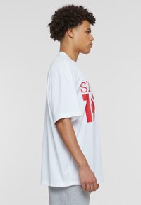 Camiseta de gran tamaño blanca con mangas cortas, que presenta texto en negrita rojo y el número '75' en la parte delantera. Material de algodón, textura suave.