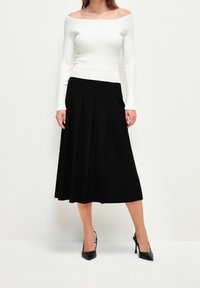 Schwarzer plissierter Rock mit einem taillierten Bund, kombiniert mit einem gerippten weißen Off-Shoulder-Top. Das Model trägt schwarze High-Heels.