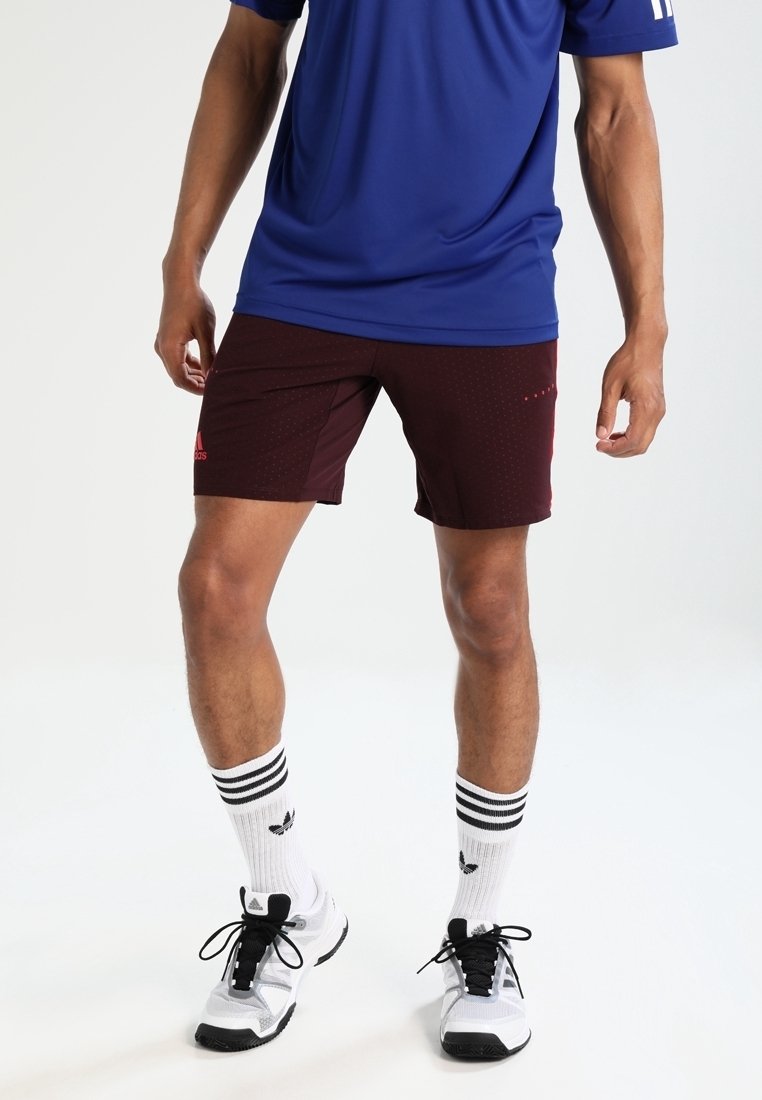 Bordeauxfärgade atletiska shorts med prickigt mönster, röd logotypdetalj, tillsammans med en blå t-shirt. Vita strumpor med svarta ränder och svarta sneakers.