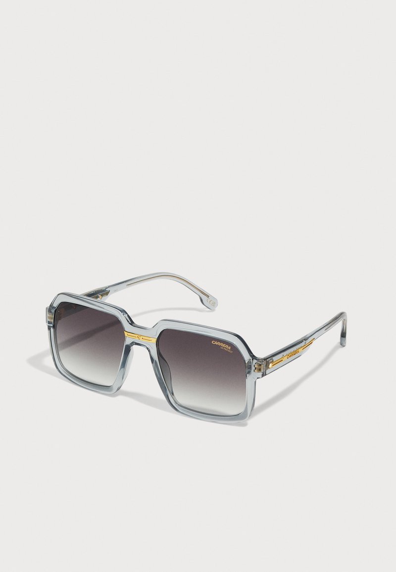 Occhiali da sole rettangolari grigi con lenti sfumate, montatura trasparente e dettagli dorati sulle tempie. Presentano un design audace e geometrico.