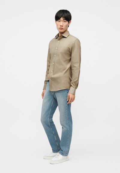Νεαρός άνδρας στέκεται φορώντας μια μπεζ πουκάμισο με κουμπιά, ανοιχτόχρωμο τζιν και λευκά αθλητικά παπούτσια, μπροστά σε ένα απλό λευκό φόντο.