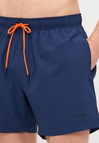 Námořnické plavky s elastickým pasem, oranžovou šňůrkou, bočními kapsami a černým logem na levé straně. Hladká textilie.