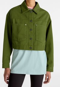 Giacca di jeans verde oliva tagliata, con due tasche frontali e bottoni neri, indossata sopra una t-shirt azzurro chiaro, abbinata a leggings neri.