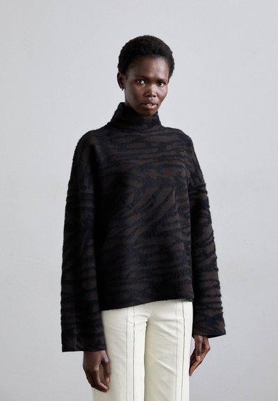 Pull-over noir surdimensionné avec une texture douce et duveteuse, présentant un motif zébré et un col montant, associé à un pantalon blanc.