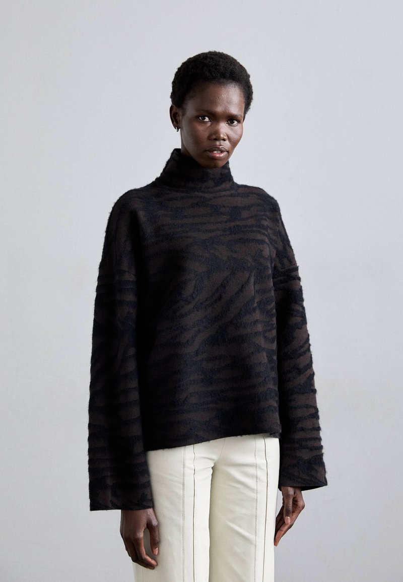 Suéter negro de gran tamaño con una textura suave y peluda, con un patrón de rayas de tigre y un cuello alto, combinado con pantalones blancos.
