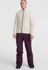 Veste en polaire gris clair avec une fermeture éclair noire, portée avec un pantalon cargo violet foncé avec poches latérales et coupe décontractée. Chaussures noires.