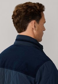 Donkere marineblauwe gewatteerde jas met een gebreide corduroy schouderdeel. Voorzien van een hoge kraag en zichtbare naaddetails, wat een gelaagde uitstraling geeft.