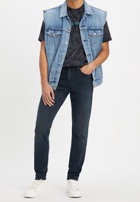 Chaleco de denim con un corte clásico, color azul desgastado, cuatro bolsillos, superpuesto a una camiseta negra de tie-dye; combinado con jeans oscuros y zapatillas blancas.