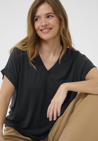 Juodas V-apskritimo marškinėliai su suktomis trumpomis rankovėmis, pagaminti iš minkštos medžiagos, dėvimi su bežinėmis kelnėmis. Modelis sėdi atsipalaidavusioje pozicijoje.