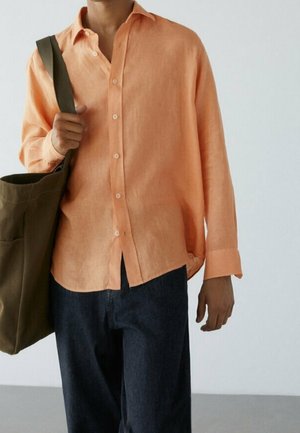 Person wearing a loose peach button-up shirt and dark pants, carrying a large olive-green shoulder bag.