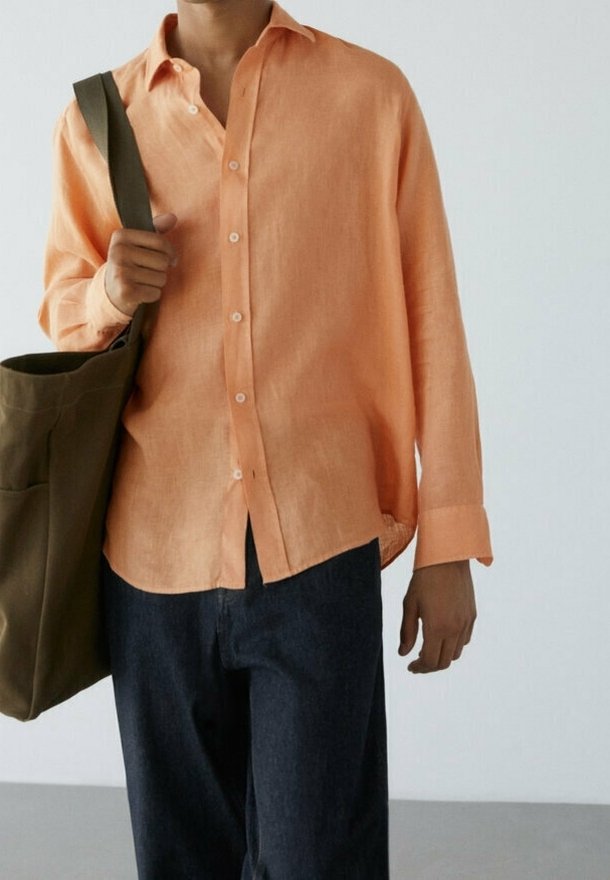 Personne portant une chemise ample couleur pêche à boutons et un pantalon foncé, portant un grand sac à bandoulière vert olive.
