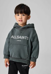 Felpa con cappuccio in pile verde scuro con tasca frontale, con la scritta "ALLSAINTS LONDON" in bianco. Indossata con pantaloni neri con logo.