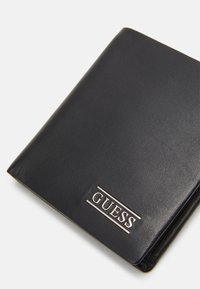 Portafoglio in pelle nera con superficie testurizzata, caratterizzato da un accentuato logo "GUESS" in argento sulla parte anteriore. Forma rettangolare compatta.