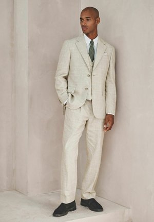 Man in beige plaid three-piece suit with green tie and black shoes standing against a light gray wall.