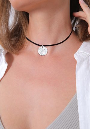 Choker noir en tissu texturé, avec un pendentif rond en argent au fini brossé, porté autour du cou.